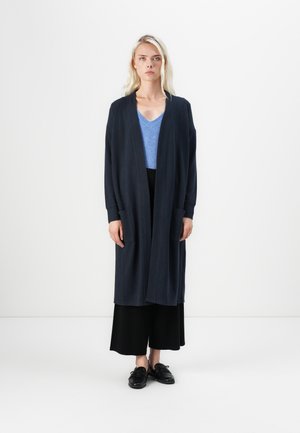 Lang marineblå cardigan med åpen front og to frontlommer, brukt over en lyseblå v-hals topp og svarte vide bukser. Svarte sko.