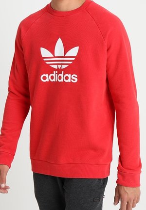 Rotes Adidas-Sweatshirt mit weißem Trefoil-Logo, getragen von einer Person vor einem einfarbigen hellen Hintergrund.