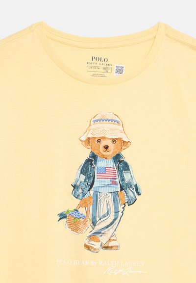 Μπλουζάκι με αρκούδο που φοράει καπέλο, τζιν μπουφάν, ριγέ παντελόνι και κρατάει καλάθι με μπλε σταφύλια, σε ανοιχτό κίτρινο φόντο.