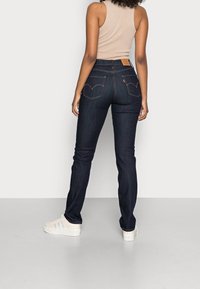 Dark blue denim jeans featuring a slim fit, gold stitching, and classic back pockets with a curved design. Paired with a beige tank top.