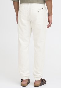 Vue arrière d'une personne portant un pantalon de jogging blanc avec des poches arrière boutonnées et des sandales marron sur un fond blanc.