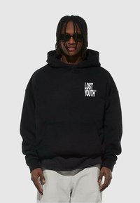 Svart hoodie i mjukt material, med en frontficka, dragsko i huvan och vit text "LOST YOUTH" på vänster bröstområde.