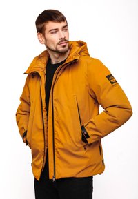 Giacca isolante arancione con cappuccio, zip completa, tasche laterali, accenti neri e patch con logo sulla manica. Superficie esterna liscia.