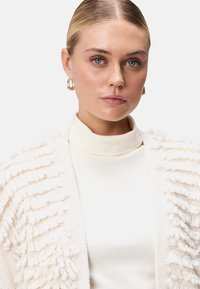 Crème-kleurige gebreide cardigan met getextureerde franjes, gedragen over een bijpassend turtleneck top. Kenmerkt zich door een soepele stof en grote ringoorbellen.