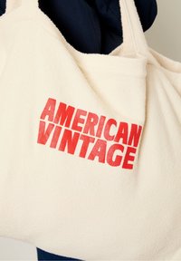 Sac fourre-tout en polaire couleur crème avec des poignées épaisses, affichant un texte rouge vif "AMERICAN VINTAGE" imprimé sur un côté. Texture douce, forme arrondie.