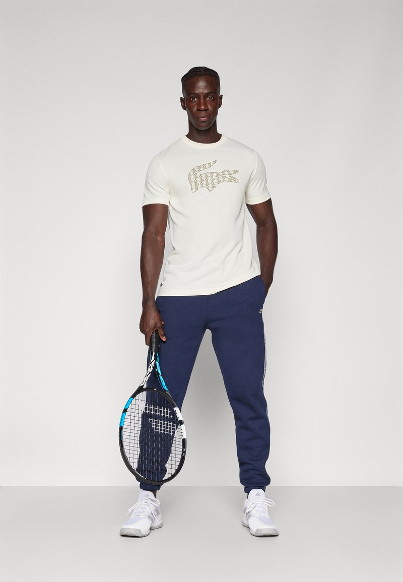 T-shirt crème avec un logo crocodile doré, associé à un pantalon de jogging bleu marine. Tenant une raquette de tennis ; chaussures de sport blanches. Design simple et sportif.