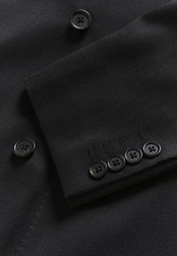 Juodas pritaikytas švarkas su briaunotu tekstūru, dviem dideliais priekiniais mygtukais ir rankove, kuri turi keturis mažesnius suderintus mygtukus.