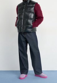 Person som bär en svart pufferväst, en burgundy fluffig tröja, vida mörka jeans och lavendelfärgade skor med rosa sula, stående på en blå golv.