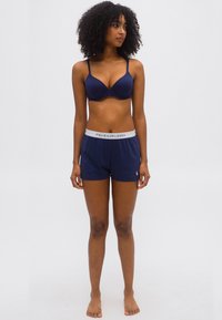 Γυναίκα με σγουρά μαλλιά, φορώντας μπλε σουτιέν και αντίστοιχα σορτς Polo Ralph Lauren, όρθια ξυπόλητη μπροστά σε λευκό απλό φόντο.