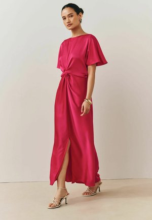 Frau trägt ein leuchtend pinkfarbenes, kurzärmeliges Kleid mit Knotendetail vorne und Schlitz, kombiniert mit weißen, schmalen Sandalen mit Absatz, stehend im Innenraum.