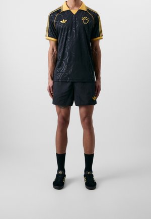 Mann trägt schwarze und gelbe Adidas-Sportbekleidung, darunter ein Shirt, Shorts, Socken und Sneakers, steht vor einem einfarbigen grauen Hintergrund.