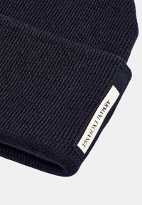 Berretto lavorato a maglia di colore blu navy con una texture a coste. Presenta un'etichetta bianca con la scritta "ARANYA EXCHANGE" sul lato. Materiale liscio e morbido.