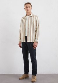 Gestreepte shirt in beige, groen en wit, met een knopenfront, borstzak en een zachte textuur; gecombineerd met marineblauwe broeken en bruine schoenen.