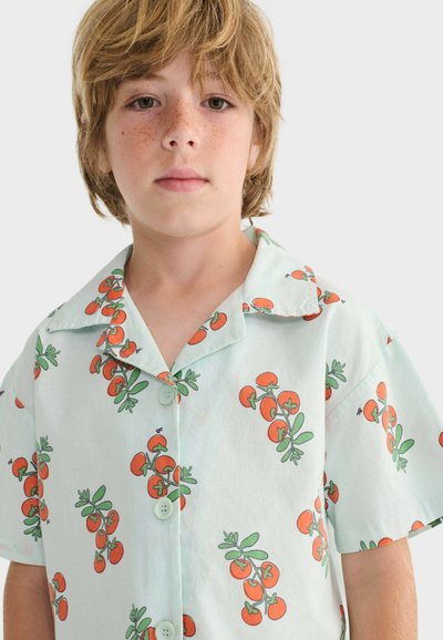 Enfant blond avec des taches de rousseur portant une chemise à manches courtes bleu clair ornée de motifs de tomates cerises rouges et de tiges vertes, debout devant un fond uni.