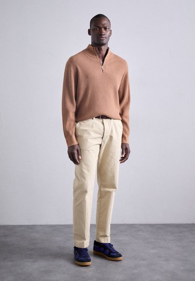 Brun ribbet genser med halv glidelås, lysebeige bukser og blå joggesko med mørke aksenter. Modellen står mot en nøytral bakgrunn.