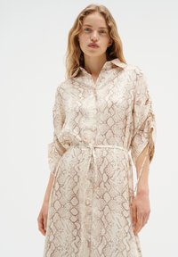 Pitkä, kevyt paitamekko kerman värissä, jossa on ruskea käärmeennahkakuosi. Ominaisuuksina kaulus, nappikansi ja vyötärösolki. Löysät hihat, joissa on kiristysnauhat.