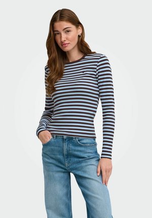 Jeune femme aux longs cheveux bruns portant un t-shirt à manches longues rayé bleu et marron et un jean bleu clair avec une main dans la poche.