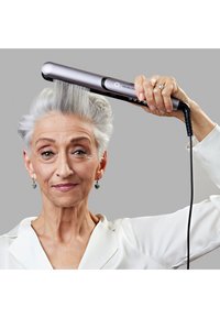 Piastra per capelli argentata dal design elegante con dettagli neri, tenuta sopra i capelli grigi acconciati, su uno sfondo grigio. Presenta un manico ergonomico e un cavo.