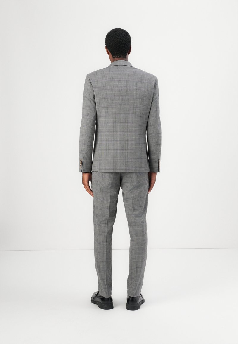 Grijze geruite pak met een getailleerde pasvorm, voorzien van een blazer met twee knopen en rechte broeken. Afgemaakt met zwarte schoenen.