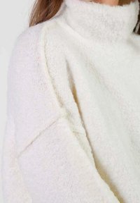 Miękki, biały sweter z golfem o puszystej teksturze noszony przez osobę z długimi brązowymi włosami, eksponujący detale na ramionach i rękawach.