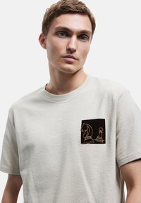 Camiseta de manga corta en gris claro, fabricada en un tejido texturizado, con un parche cuadrado marrón oscuro que presenta piezas de ajedrez bordadas. Cuello redondo.