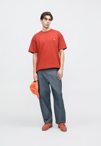 Κόκκινο βαμβακερό μπλουζάκι με κοντά μανίκια και λογότυπο, συνδυασμένο με φαρδιά μπλε τζιν παντελόνια και πορτοκαλί καπέλο, κρατημένο στο αριστερό χέρι.