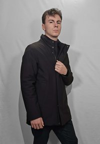 Giacca nera, isolante, con collo alto e bottoni a pressione. Presenta una trama liscia e tasche laterali, indossata sopra una maglia nera e jeans.