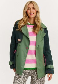 Zeleno-námořnický kabát s širokým límcem, růžovými akcenty a dvouřadým zapínáním, kombinovaný s pruhovaným svetrem a kalhotami s leopardím vzorem.