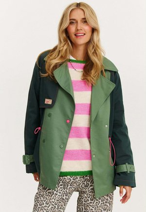 Zielono-granatowa kurtka z szerokim kołnierzem, różowymi akcentami i podwójnym zapięciem na guziki, zestawiona z paskowanym swetrem i spodniami w panterkę.