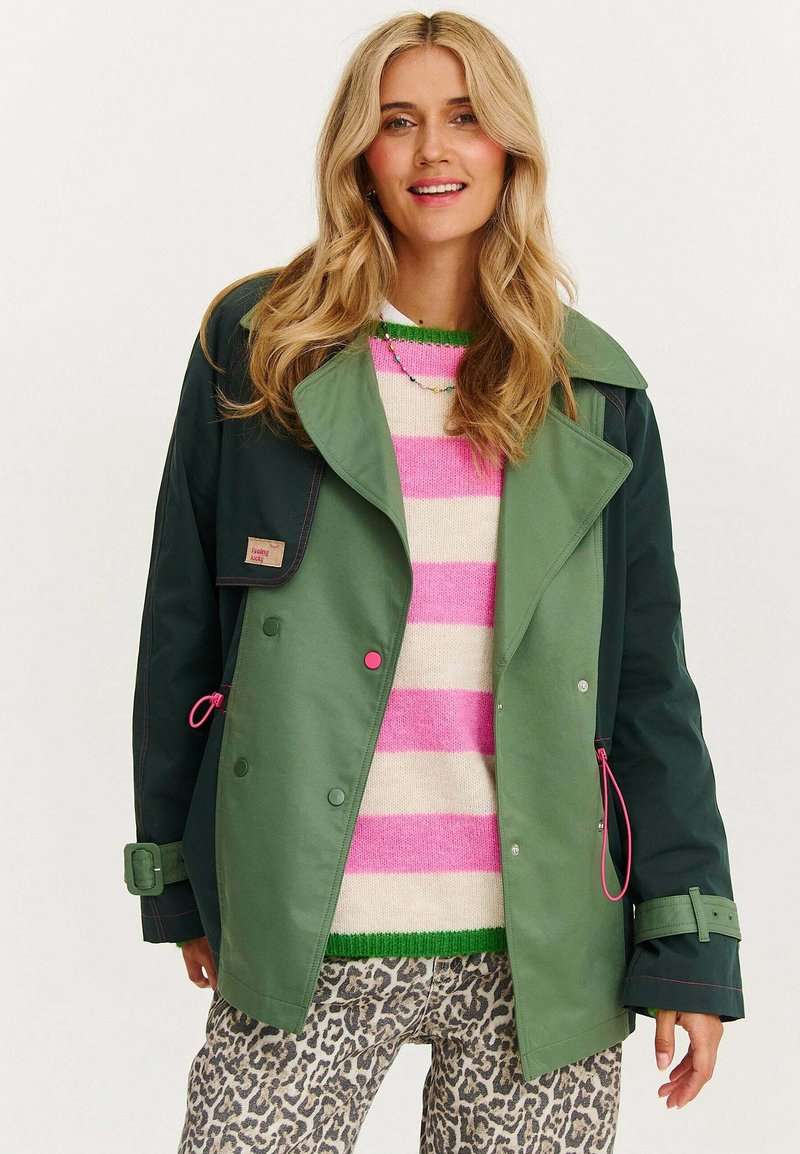 Zeleno-námořnický kabát s širokým límcem, růžovými akcenty a dvouřadým zapínáním, kombinovaný s pruhovaným svetrem a kalhotami s leopardím vzorem.