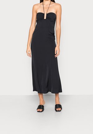 Czarna bezramiankowa sukienka midi z dekoltem w kształcie serca i cienkim paskiem na szyję, noszona z czarnymi wsuwanymi sandałami z wyściełaną podeszwą na jasnym tle.