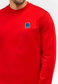 Camicia a maniche lunghe rossa in tessuto liscio. Presenta un logo rettangolare blu sulla parte sinistra del petto. Design classico a girocollo.