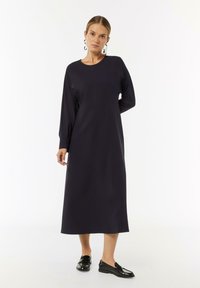 Langes, dunkelblaues Kleid mit rundem Ausschnitt, langen Ärmeln und gerader Silhouette. Getragen mit schwarzen Slippern, die eine glatte Textur aufweisen.