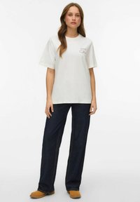 Biała bawełniana koszulka z krótkim rękawem, z małym nadrukiem na górnym lewym rogu. Połączona z szerokimi ciemnoniebieskimi dżinsami i jasnobrązowym obuwiem.