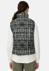 Vrouw met lang bruin haar in een paardenstaart, gekleed in een zwart-wit gestreepte puffer vest over een wit langemouwen shirt en olijfgroene broek.
