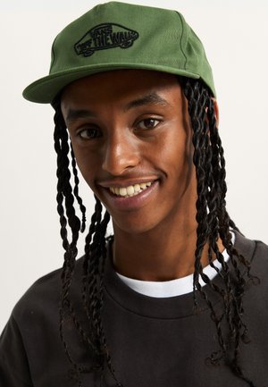 Gorra verde con visera plana y logotipo negro bordado. La persona lleva el cabello largo trenzado y una sudadera oscura con un cuello blanco.