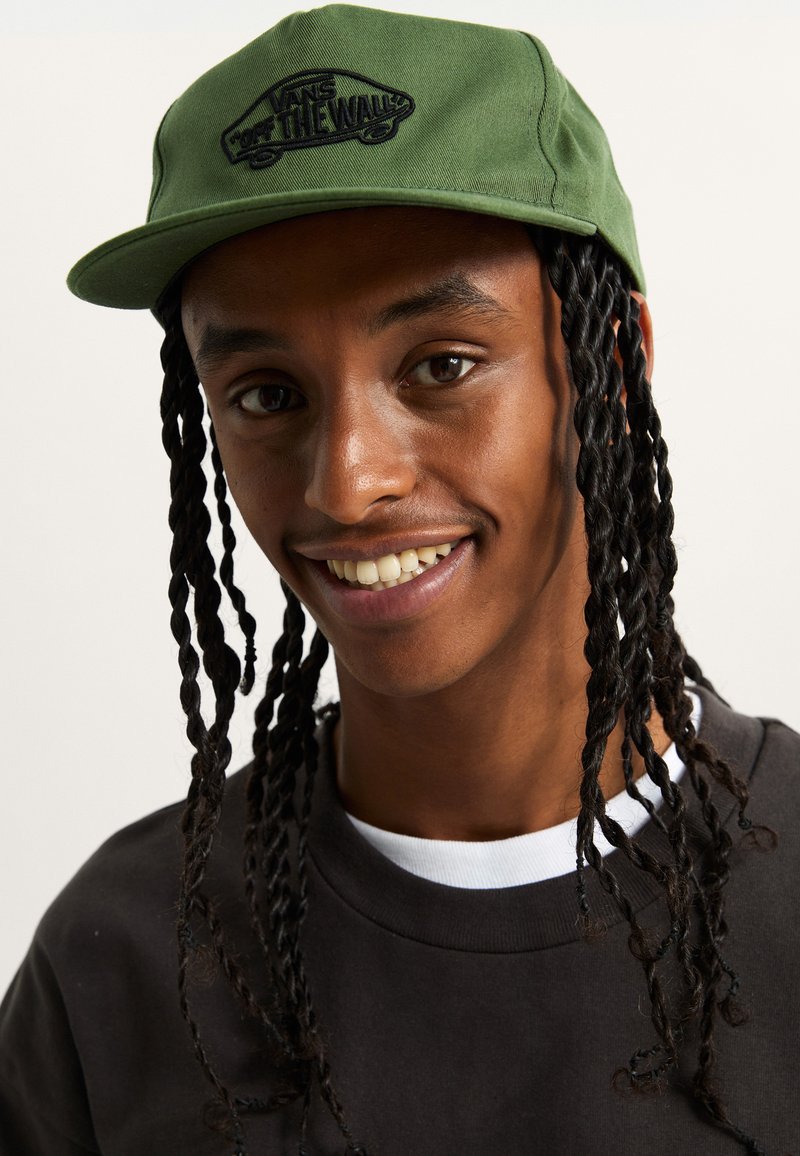 Grüne Kappe mit flachem Schild und schwarzem gesticktem Logo. Der Träger hat lange geflochtene Haare und trägt einen dunklen Pullover mit weißem Kragen.