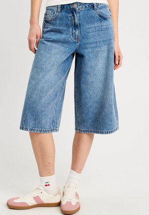 Person iført mid-lange blå denimshorts med forlommer, hvide sokker med kirsebærmotiv og hvide og lyserøde sneakers.