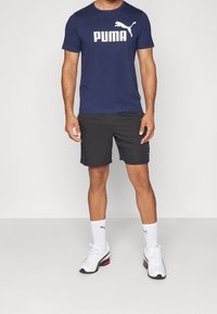Hombre vistiendo una camiseta navy de Puma, pantalones cortos negros, calcetines blancos de Puma y zapatillas blancas, de pie sobre un fondo liso.