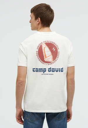 Man wearing white T-shirt with orange and blue windsurfing graphic and text on back, paired with blue jeans, facing sideways.