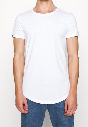 Mand iført en ensfarvet hvid kortærmet t-shirt og blå jeans, stående mod en lys, tom baggrund med armene afslappet langs siden.