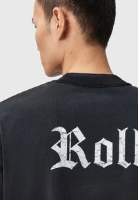 Czarna bawełniana koszulka z dużym, popękanym białym napisem "Roll" na plecach, okrągłym dekoltem i widocznymi detalami szwów.