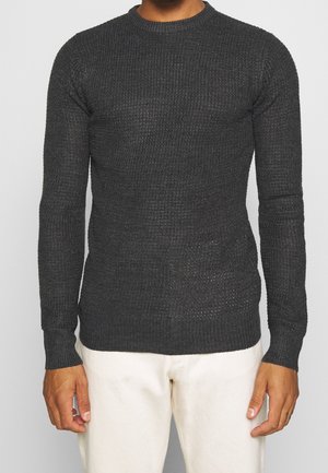 Pull en maille gris foncé avec un col rond, des manches longues et une finition texturée. Porté avec un pantalon de couleur claire.
