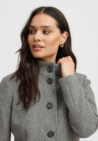 Manteau en laine gris avec un col haut, quatre boutons noirs à l'avant et une finition texturée douce. Gros plan sur la zone du cou et des épaules.