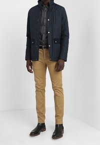 Chaqueta acolchada navy con botones a presión dorados, camisa a cuadros en blanco y negro, chinos beige y botas de piel negras hasta el tobillo.