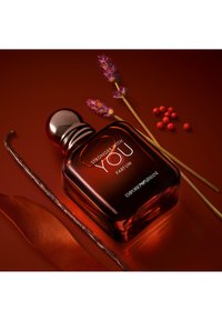 Flacone di profumo ambra scuro Emporio Armani "Stronger With You" con rametti di lavanda, bacche rosse, baccello di vaniglia e foglia marrone su una superficie marrone.