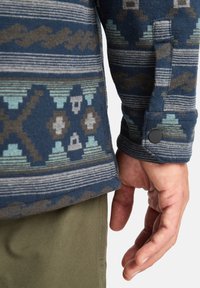 Navy patterned fleece jacket with geometric designs in teal, gray, and black. Features a buttoned cuff and horizontal stripe detailing.