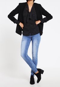 Schwarze Blazer über einer schwarz-weißen Bluse mit Punkten und einer Schleife. Helle, verwaschene Skinny-Jeans in Blau und schwarze, blockabsätzige Stiefeletten.