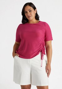 Top fucsia a maniche corte con dettagli arricciati sui lati, realizzato in un tessuto lucido e testurizzato; abbinato a pantaloni corti bianchi fino al ginocchio.