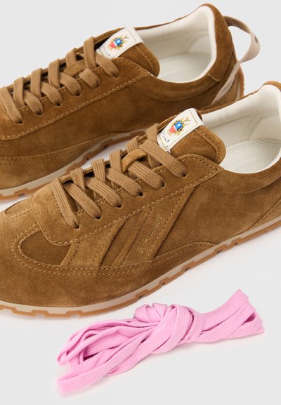 Ténis de camurça castanhos com interior creme, sola de borracha bege e atacadores castanhos. Inclui atacadores cor-de-rosa e um rótulo com um brasão.